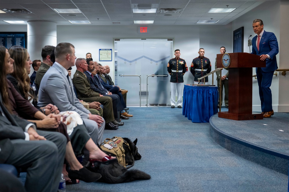 A formal ceremony or briefing is taking place in what appears to be a government or military facility, likely the Pentagon, indicated by the Department of Defense seal visible on the podium. A man in a blue suit is speaking at a wooden lectern while an audience of military personnel in uniform and civilians seated in rows listens attentively. Two U.S. Marines in dress uniform stand at attention near the entrance, and a military working dog with a tactical vest is visible among the seated attendees.