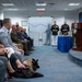 A formal ceremony or briefing is taking place in what appears to be a government or military facility, likely the Pentagon, indicated by the Department of Defense seal visible on the podium. A man in a blue suit is speaking at a wooden lectern while an audience of military personnel in uniform and civilians seated in rows listens attentively. Two U.S. Marines in dress uniform stand at attention near the entrance, and a military working dog with a tactical vest is visible among the seated attendees.