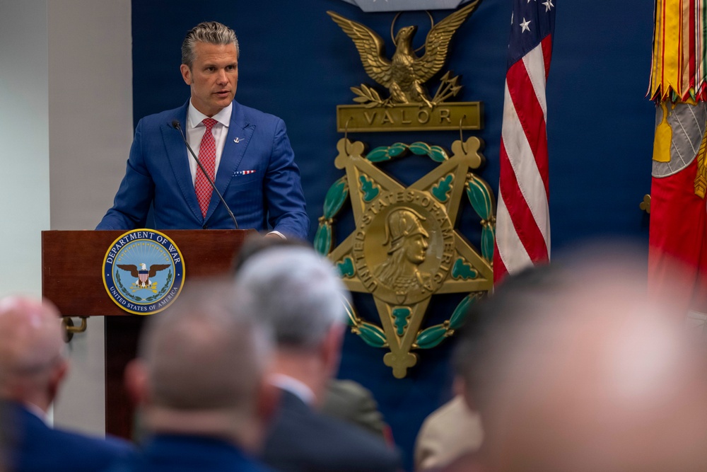 A man in a blue suit with a red tie is speaking at a podium bearing the seal of the 'Department of War, United States of America' before an audience. Behind him is a large Medal of Honor emblem with the word 'VALOR' and an American flag alongside military unit colors. The setting appears to be a formal military or government ceremony.