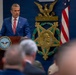 A man in a blue suit with a red tie is speaking at a podium bearing the seal of the 'Department of War, United States of America' before an audience. Behind him is a large Medal of Honor emblem with the word 'VALOR' and an American flag alongside military unit colors. The setting appears to be a formal military or government ceremony.