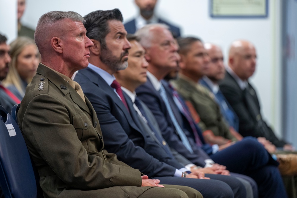 A formal gathering showing a row of seated attendees, including a U.S. Marine Corps officer in dress uniform prominently in the foreground alongside several civilians in business attire. The group appears to be attending an official ceremony, briefing, or formal event, all facing the same direction as if watching a presentation or speaker. The setting appears to be an indoor institutional or government facility.