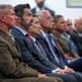 A formal gathering showing a row of seated attendees, including a U.S. Marine Corps officer in dress uniform prominently in the foreground alongside several civilians in business attire. The group appears to be attending an official ceremony, briefing, or formal event, all facing the same direction as if watching a presentation or speaker. The setting appears to be an indoor institutional or government facility.