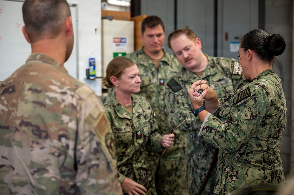A group of military personnel in various camouflage uniforms are gathered in what appears to be an indoor training or workshop environment. A female service member on the right, wearing Navy Working Uniform (NWU Type III), appears to be demonstrating or instructing others using a small object or tool. The group includes personnel wearing both Army Operational Camouflage Pattern (OCP) and Navy/Marine Corps digital camouflage uniforms.