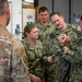 A group of military personnel in various camouflage uniforms are gathered in what appears to be an indoor training or workshop environment. A female service member on the right, wearing Navy Working Uniform (NWU Type III), appears to be demonstrating or instructing others using a small object or tool. The group includes personnel wearing both Army Operational Camouflage Pattern (OCP) and Navy/Marine Corps digital camouflage uniforms.