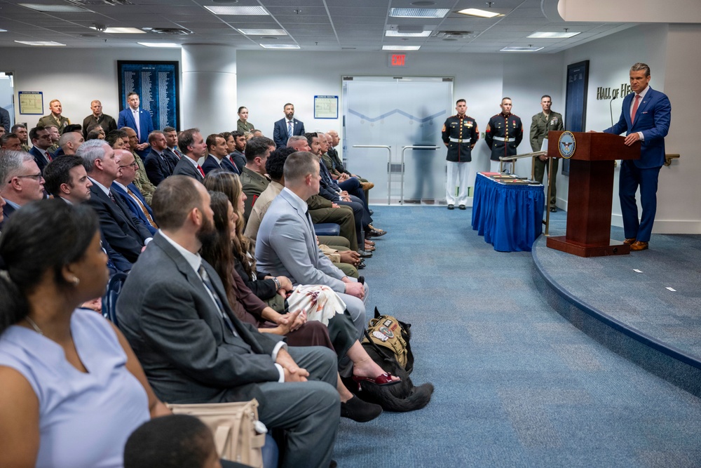 A formal ceremony is taking place in what appears to be the Pentagon's Hall of Heroes, as indicated by signage visible on the right wall. A speaker in a blue suit addresses a seated audience of mixed civilians and military personnel from a wooden podium bearing the Department of Defense seal. Three U.S. Marines in dress uniforms stand at attention near the back of the room.