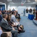 A formal ceremony is taking place in what appears to be the Pentagon's Hall of Heroes, as indicated by signage visible on the right wall. A speaker in a blue suit addresses a seated audience of mixed civilians and military personnel from a wooden podium bearing the Department of Defense seal. Three U.S. Marines in dress uniforms stand at attention near the back of the room.
