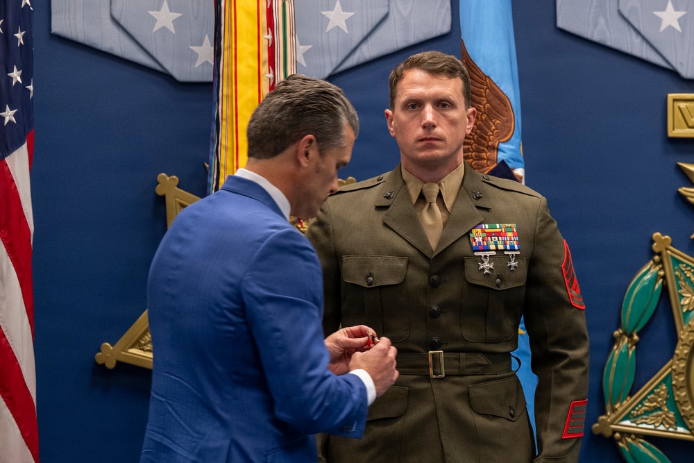 A formal military awards ceremony is taking place, likely at the Pentagon based on the distinctive blue wall decor and military seals visible in the background. A man in a blue civilian suit is pinning a medal or award onto the uniform of a U.S. Marine Corps Staff Sergeant or Gunnery Sergeant in dress olive uniform. The Marine stands at attention with existing ribbons and medals already displayed on his chest.