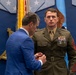A formal military awards ceremony is taking place, likely at the Pentagon based on the distinctive blue wall decor and military seals visible in the background. A man in a blue civilian suit is pinning a medal or award onto the uniform of a U.S. Marine Corps Staff Sergeant or Gunnery Sergeant in dress olive uniform. The Marine stands at attention with existing ribbons and medals already displayed on his chest.