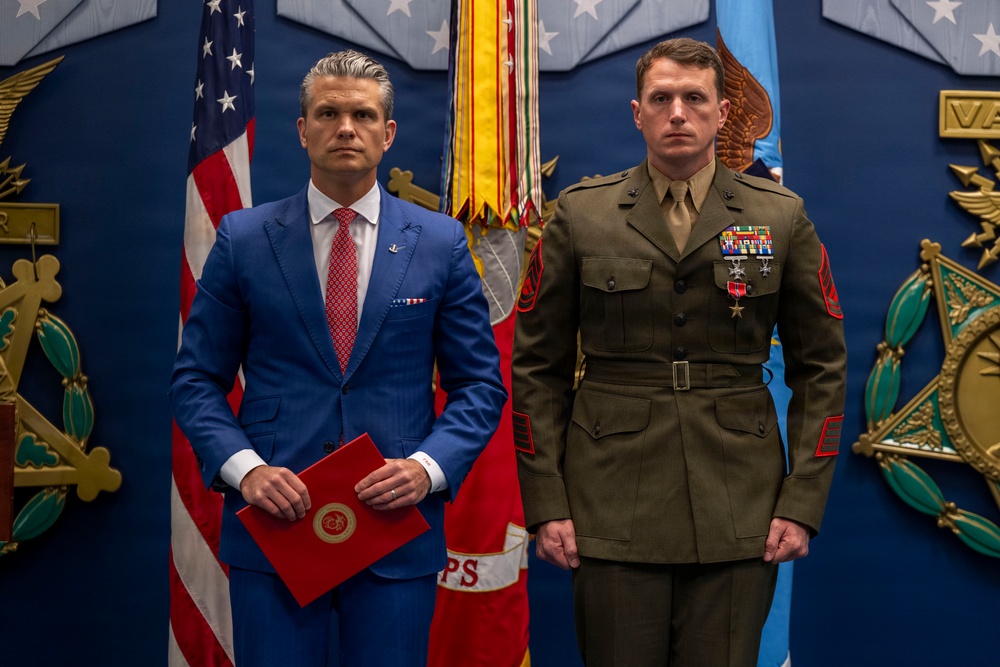 Two men stand formally in front of an American flag and military ceremonial backdrop. One man wears a blue civilian suit and holds a red folder with an official seal, while the other wears a U.S. Marine Corps dress uniform with multiple medals and ribbons. The setting appears to be an official military awards or recognition ceremony.