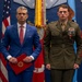 Two men stand formally in front of an American flag and military ceremonial backdrop. One man wears a blue civilian suit and holds a red folder with an official seal, while the other wears a U.S. Marine Corps dress uniform with multiple medals and ribbons. The setting appears to be an official military awards or recognition ceremony.