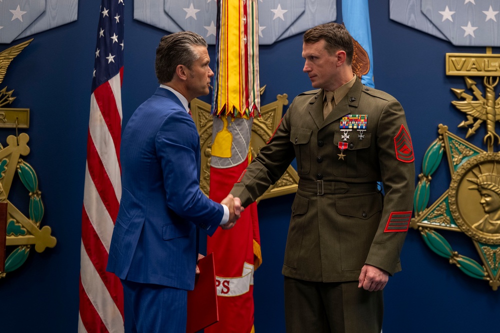 A formal military ceremony is taking place in what appears to be the Pentagon or a similar high-level defense facility. A civilian in a blue suit is shaking hands with a U.S. Marine Corps senior non-commissioned officer in dress uniform, who has recently received a medal. The setting features official military decorations, flags, and emblems on the wall.