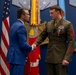 A formal military ceremony is taking place in what appears to be the Pentagon or a similar high-level defense facility. A civilian in a blue suit is shaking hands with a U.S. Marine Corps senior non-commissioned officer in dress uniform, who has recently received a medal. The setting features official military decorations, flags, and emblems on the wall.