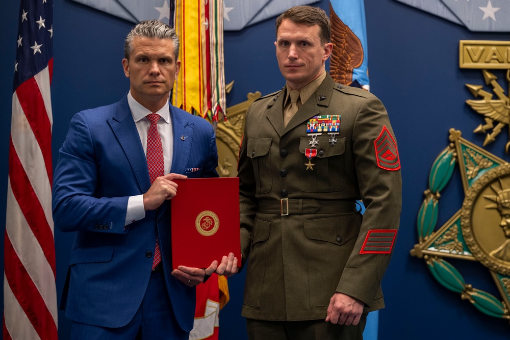 Two men pose formally in what appears to be an official military ceremony setting. A civilian in a blue suit holds a red official folder bearing a Marine Corps emblem, standing beside a U.S. Marine in dress uniform adorned with multiple medals and rank insignia. The background features the U.S. flag, military color flags, and a large decorative military emblem on a blue wall.