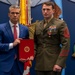 Two men pose formally in what appears to be an official military ceremony setting. A civilian in a blue suit holds a red official folder bearing a Marine Corps emblem, standing beside a U.S. Marine in dress uniform adorned with multiple medals and rank insignia. The background features the U.S. flag, military color flags, and a large decorative military emblem on a blue wall.