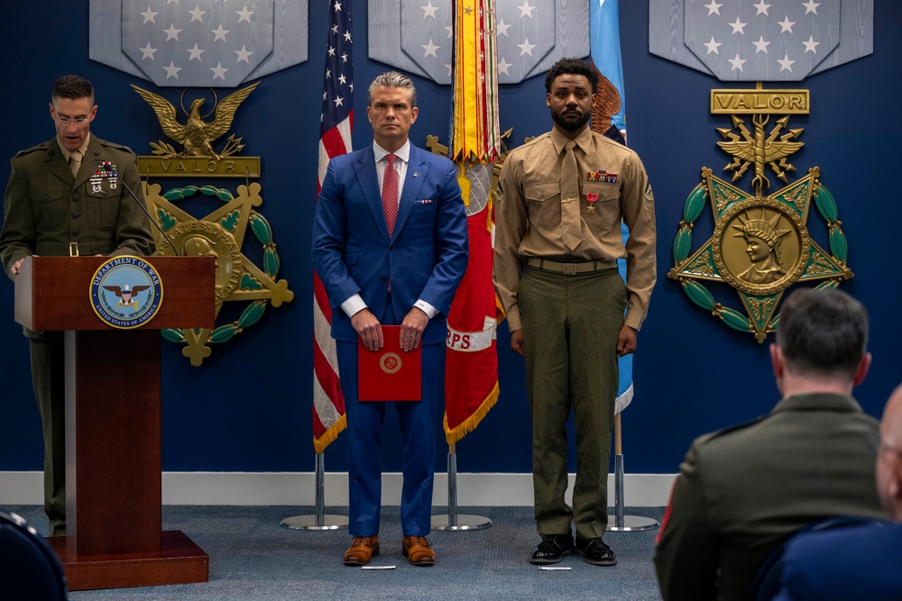 A formal military ceremony is taking place at the Pentagon or a Department of Defense facility. A Marine officer speaks at a podium while a civilian in a blue suit and a Marine in service uniform stand at attention. The setting features Medal of Honor and 'VALOR' emblems on the wall, along with military flags including the U.S. flag and Marine Corps flag.