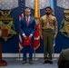 A formal military ceremony is taking place at the Pentagon or a Department of Defense facility. A Marine officer speaks at a podium while a civilian in a blue suit and a Marine in service uniform stand at attention. The setting features Medal of Honor and 'VALOR' emblems on the wall, along with military flags including the U.S. flag and Marine Corps flag.