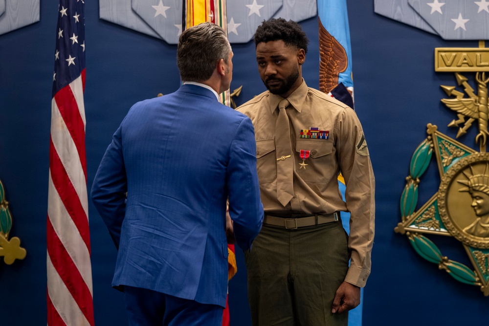 A formal military ceremony is taking place in what appears to be an official government or military building. A U.S. Marine in service uniform is receiving recognition, visible by a medal pinned to his chest, from a civilian in a blue suit. The setting includes an American flag, military insignia, and what appears to be a Medal of Honor display on the wall.