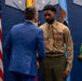 A formal military ceremony is taking place in what appears to be an official government or military building. A U.S. Marine in service uniform is receiving recognition, visible by a medal pinned to his chest, from a civilian in a blue suit. The setting includes an American flag, military insignia, and what appears to be a Medal of Honor display on the wall.