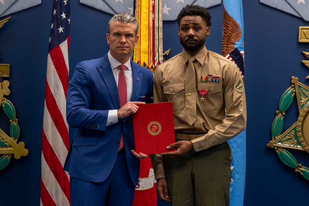 Two individuals pose formally in what appears to be an official military ceremony setting. A man in a blue suit and red tie stands alongside a U.S. Marine in service uniform (tan coat, green trousers), both holding a red folder bearing the U.S. Marine Corps emblem. The Marine is wearing several ribbons and a medal, suggesting this is an award or commendation presentation.