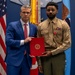 Two individuals pose formally in what appears to be an official military ceremony setting. A man in a blue suit and red tie stands alongside a U.S. Marine in service uniform (tan coat, green trousers), both holding a red folder bearing the U.S. Marine Corps emblem. The Marine is wearing several ribbons and a medal, suggesting this is an award or commendation presentation.