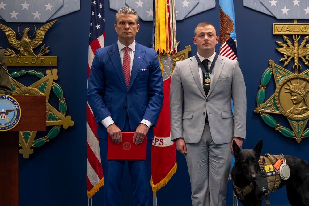 A formal military or government awards ceremony is taking place in front of a blue wall decorated with 'VALOR' insignia and large military medals. Two men in suits stand at attention, with the man on the right wearing a medal around his neck and standing beside a black service dog in a tactical vest. The setting appears to be an official Department of the Army or Department of Defense venue.