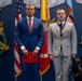 A formal military or government awards ceremony is taking place in front of a blue wall decorated with 'VALOR' insignia and large military medals. Two men in suits stand at attention, with the man on the right wearing a medal around his neck and standing beside a black service dog in a tactical vest. The setting appears to be an official Department of the Army or Department of Defense venue.