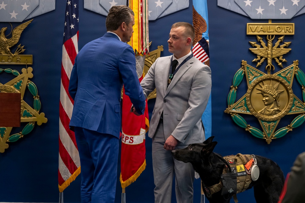 A formal military or government ceremony is taking place in front of a display wall featuring the Medal of Honor emblem and 'VALOR' plaques. Two men in suits are shaking hands, with one wearing a light gray suit accompanied by a black service dog in a tactical vest. The setting includes American flags and military unit colors, suggesting an awards or recognition ceremony.