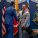 A formal military or government ceremony is taking place in front of a display wall featuring the Medal of Honor emblem and 'VALOR' plaques. Two men in suits are shaking hands, with one wearing a light gray suit accompanied by a black service dog in a tactical vest. The setting includes American flags and military unit colors, suggesting an awards or recognition ceremony.