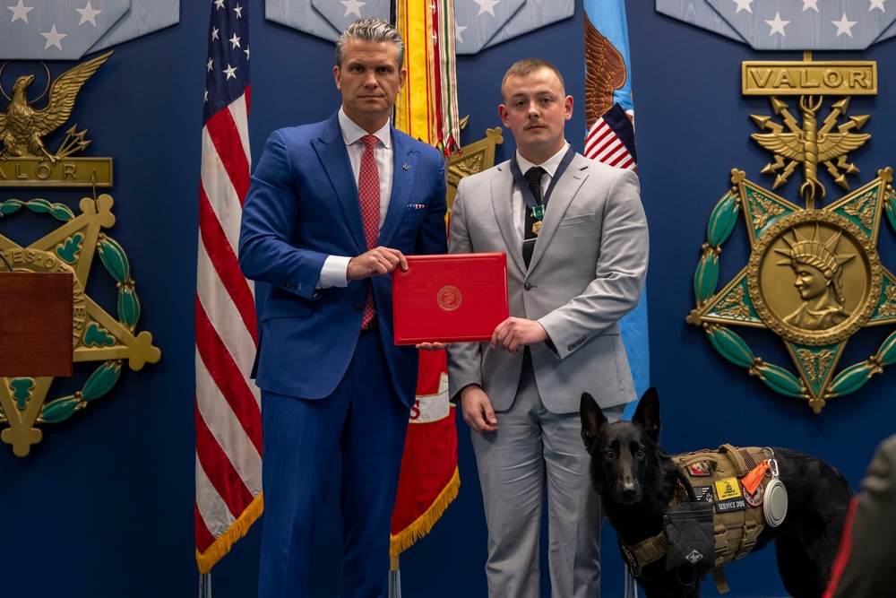 Two men are posing together in a formal military or government setting, jointly holding a red official document or award case. A black German Shepherd wearing a service dog vest stands beside them. The backdrop features prominent military insignia including Medal of Honor emblems and 'VALOR' plaques, along with an American flag.