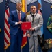 Two men are posing together in a formal military or government setting, jointly holding a red official document or award case. A black German Shepherd wearing a service dog vest stands beside them. The backdrop features prominent military insignia including Medal of Honor emblems and 'VALOR' plaques, along with an American flag.