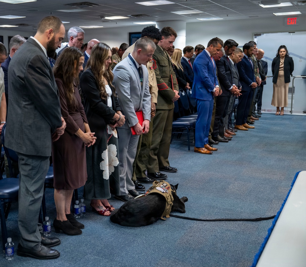 A formal indoor ceremony or memorial event with a large group of attendees standing in a line with heads bowed, suggesting a moment of silence or prayer. The group includes civilians in business attire, at least one military officer in dress uniform with medals, and a service dog wearing a tactical vest lying on the floor. The setting appears to be a government or institutional conference room.