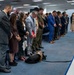 A formal indoor ceremony or memorial event with a large group of attendees standing in a line with heads bowed, suggesting a moment of silence or prayer. The group includes civilians in business attire, at least one military officer in dress uniform with medals, and a service dog wearing a tactical vest lying on the floor. The setting appears to be a government or institutional conference room.