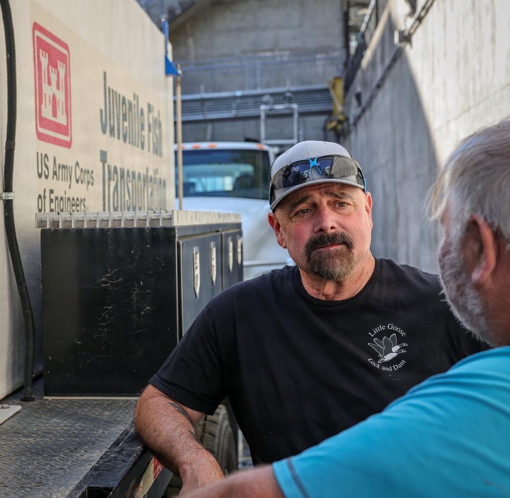 Two men are conversing near a US Army Corps of Engineers 'Juvenile Fish Transportation' vehicle at what appears to be a dam or lock facility. The man facing the camera wears a black t-shirt reading 'Little Goose Lock and Dam' and a gray cap with blue sunglasses. The setting suggests an operational fish transport or wildlife management context.