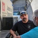 Two men are conversing near a US Army Corps of Engineers 'Juvenile Fish Transportation' vehicle at what appears to be a dam or lock facility. The man facing the camera wears a black t-shirt reading 'Little Goose Lock and Dam' and a gray cap with blue sunglasses. The setting suggests an operational fish transport or wildlife management context.