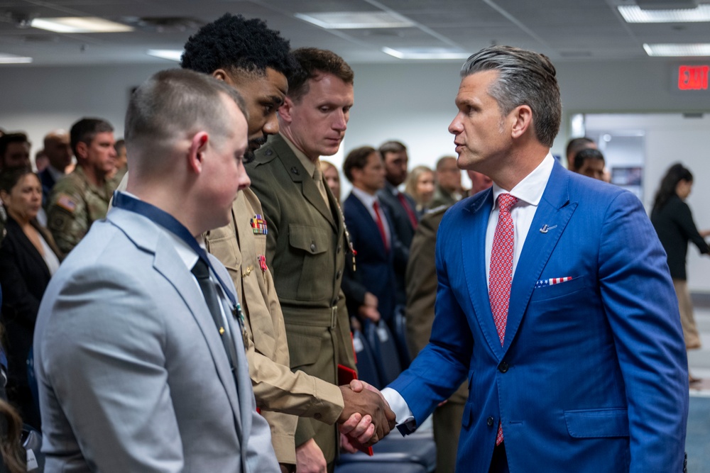A man in a blue suit with a red tie is shaking hands with a military service member in a light gray uniform. The setting appears to be an official or ceremonial gathering with multiple military personnel in various uniforms present. The event takes place in an indoor facility with civilians and uniformed personnel visible in the background.