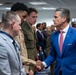 A man in a blue suit with a red tie is shaking hands with a military service member in a light gray uniform. The setting appears to be an official or ceremonial gathering with multiple military personnel in various uniforms present. The event takes place in an indoor facility with civilians and uniformed personnel visible in the background.
