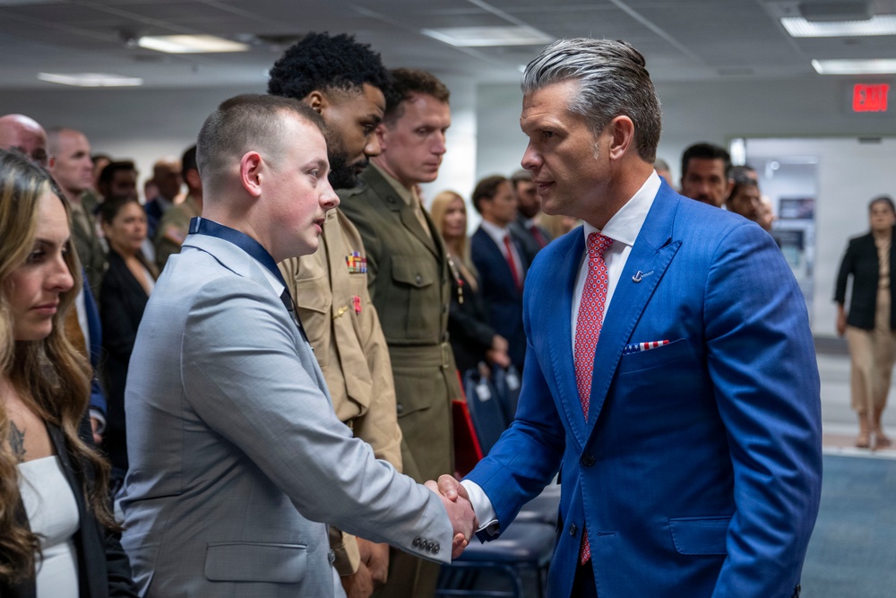 A formal indoor gathering where a man in a blue suit is shaking hands with a younger man in a gray suit. Military personnel in uniform are visible in the background, suggesting this is an official or ceremonial event. The setting appears to be a government or institutional building based on the interior design and EXIT sign visible.