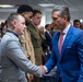 A formal indoor gathering where a man in a blue suit is shaking hands with a younger man in a gray suit. Military personnel in uniform are visible in the background, suggesting this is an official or ceremonial event. The setting appears to be a government or institutional building based on the interior design and EXIT sign visible.