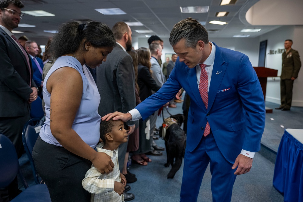 A formal indoor gathering, likely a military or government ceremony, where a man in a bright blue suit is interacting warmly with a woman and a young child by placing his hand on the child's head. The setting appears to be an official room with blue chairs, a podium, and a uniformed military officer standing in the background. A black service dog is also visible in the scene.