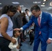 A formal indoor gathering, likely a military or government ceremony, where a man in a bright blue suit is interacting warmly with a woman and a young child by placing his hand on the child's head. The setting appears to be an official room with blue chairs, a podium, and a uniformed military officer standing in the background. A black service dog is also visible in the scene.