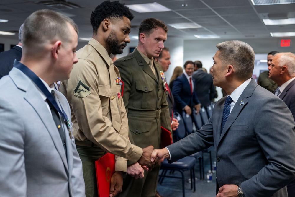 A formal indoor gathering where a civilian in a gray suit is shaking hands with a U.S. Marine in tan service uniform (Charlie uniform). Several other military personnel in various uniforms are present in the background, suggesting an official ceremony or reception. The setting appears to be a government or military facility based on the fluorescent lighting, drop ceiling, and EXIT sign visible.