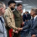 A formal indoor gathering where a civilian in a gray suit is shaking hands with a U.S. Marine in tan service uniform (Charlie uniform). Several other military personnel in various uniforms are present in the background, suggesting an official ceremony or reception. The setting appears to be a government or military facility based on the fluorescent lighting, drop ceiling, and EXIT sign visible.