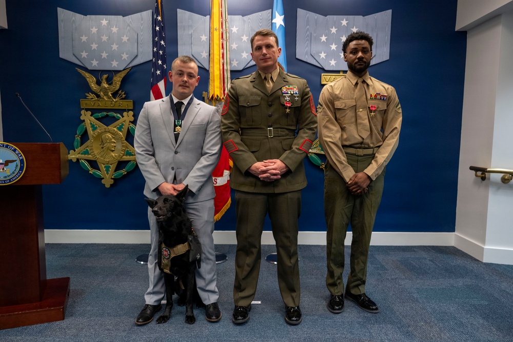 Three individuals stand posed for a formal photo in what appears to be a military or government ceremonial room, likely the Pentagon or a Department of Defense facility. Two are in military uniforms (appearing to be U.S. Marine Corps dress uniforms) and one is in a civilian suit, all wearing medals or decorations. A black service dog wearing a vest is present beside the civilian.