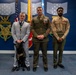 Three individuals stand posed for a formal photo in what appears to be a military or government ceremonial room, likely the Pentagon or a Department of Defense facility. Two are in military uniforms (appearing to be U.S. Marine Corps dress uniforms) and one is in a civilian suit, all wearing medals or decorations. A black service dog wearing a vest is present beside the civilian.