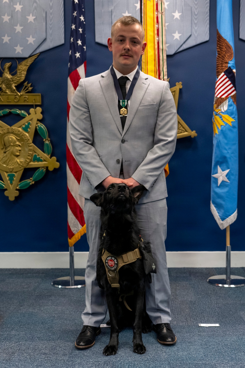 A man in a light gray suit wearing a military medal on a ribbon stands in a formal ceremonial room with a black service dog seated in front of him. The setting appears to be an official military or government building, with an American flag, a blue Air Force flag, a Medal of Honor display plaque on the wall, and decorative military pennants visible in the background. The black dog is wearing a tan service vest with patches.