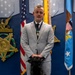 A man in a light gray suit wearing a military medal on a ribbon stands in a formal ceremonial room with a black service dog seated in front of him. The setting appears to be an official military or government building, with an American flag, a blue Air Force flag, a Medal of Honor display plaque on the wall, and decorative military pennants visible in the background. The black dog is wearing a tan service vest with patches.