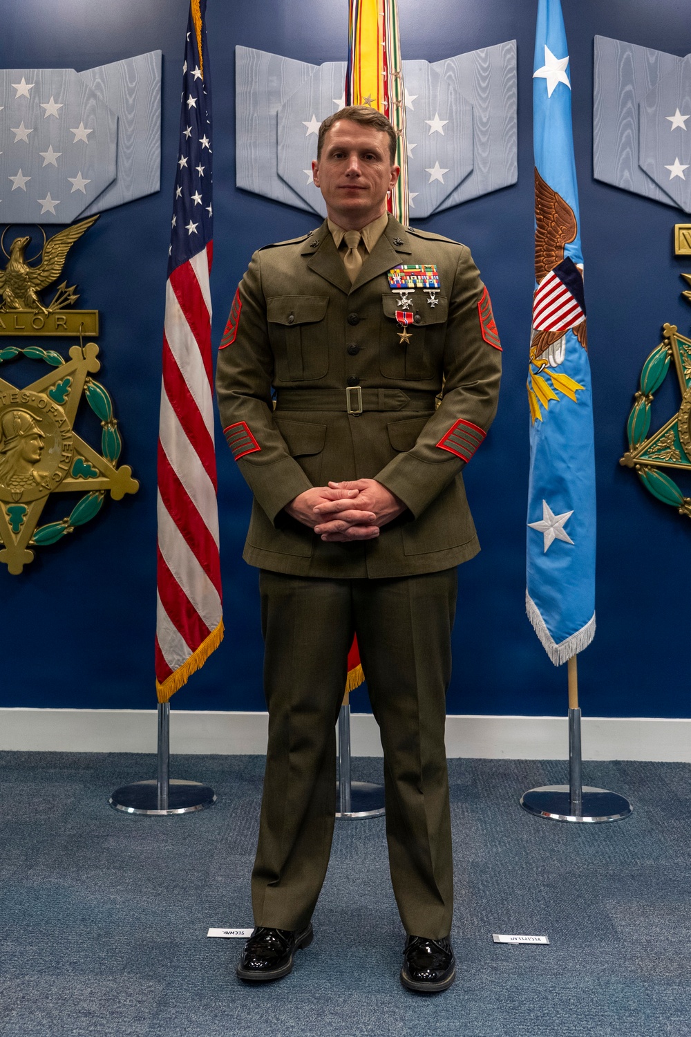 A U.S. Marine Corps staff non-commissioned officer stands at attention in dress uniform (Service 'A' greens) in what appears to be a formal Pentagon or high-level military headquarters setting. The individual is wearing multiple decorations including what appears to be a Bronze Star with valor device and additional service medals. The backdrop features Medal of Honor plaques, an American flag, and what appears to be an Air Force flag.