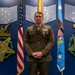A U.S. Marine Corps staff non-commissioned officer stands at attention in dress uniform (Service 'A' greens) in what appears to be a formal Pentagon or high-level military headquarters setting. The individual is wearing multiple decorations including what appears to be a Bronze Star with valor device and additional service medals. The backdrop features Medal of Honor plaques, an American flag, and what appears to be an Air Force flag.