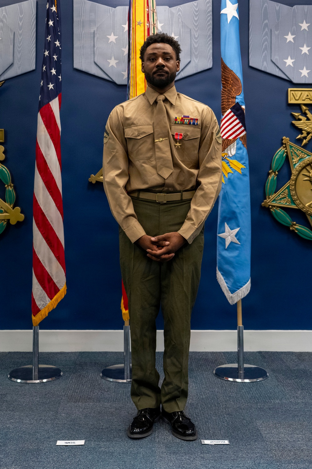 A U.S. Marine stands at attention in a formal ceremonial setting, wearing the Charlie Service uniform (khaki shirt, olive green trousers) with ribbons and what appears to be a Bronze Star Medal on his chest. He is positioned between an American flag and what appears to be an Air Force flag, in front of a blue wall decorated with military insignia and stars. The setting appears to be an official military awards or recognition ceremony.