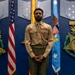 A U.S. Marine stands at attention in a formal ceremonial setting, wearing the Charlie Service uniform (khaki shirt, olive green trousers) with ribbons and what appears to be a Bronze Star Medal on his chest. He is positioned between an American flag and what appears to be an Air Force flag, in front of a blue wall decorated with military insignia and stars. The setting appears to be an official military awards or recognition ceremony.